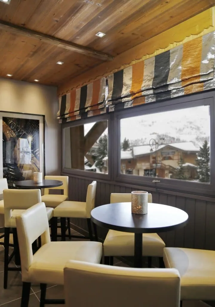 Window corner with round tables, ivory chairs and orange striped blinds overlooking snowy chalets