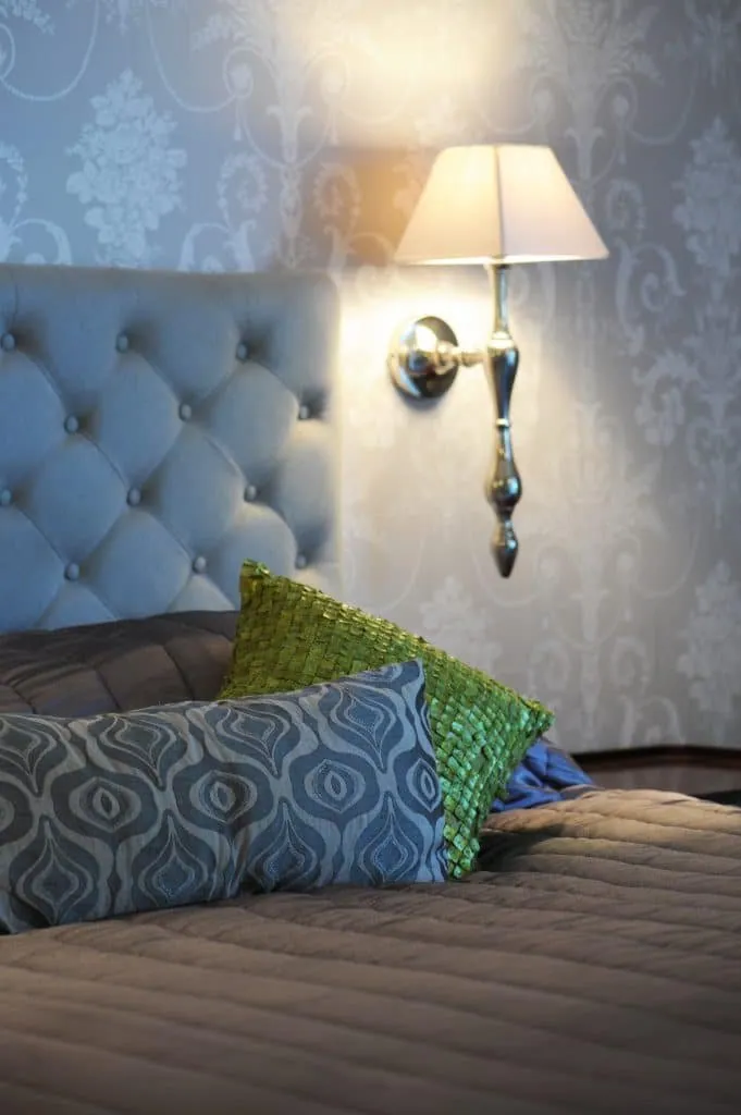 Bedroom with damask wallpaper, blue-grey buttoned headboard, silver wall light and textured green cushion