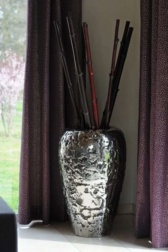 Tall textured silver vase filled with bamboo canes, between two plum curtains