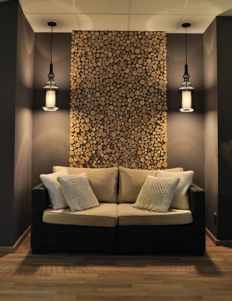 Woven sofa with cream cushions beneath a wall panel of wood slices, between two pendant lights