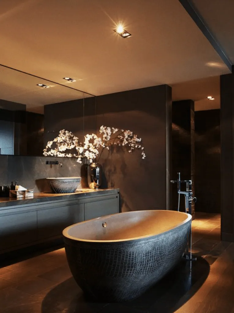 Dark bathroom under recessed spotlights: freestanding crocodile-textured bathtub, vessel basin and blossom branches