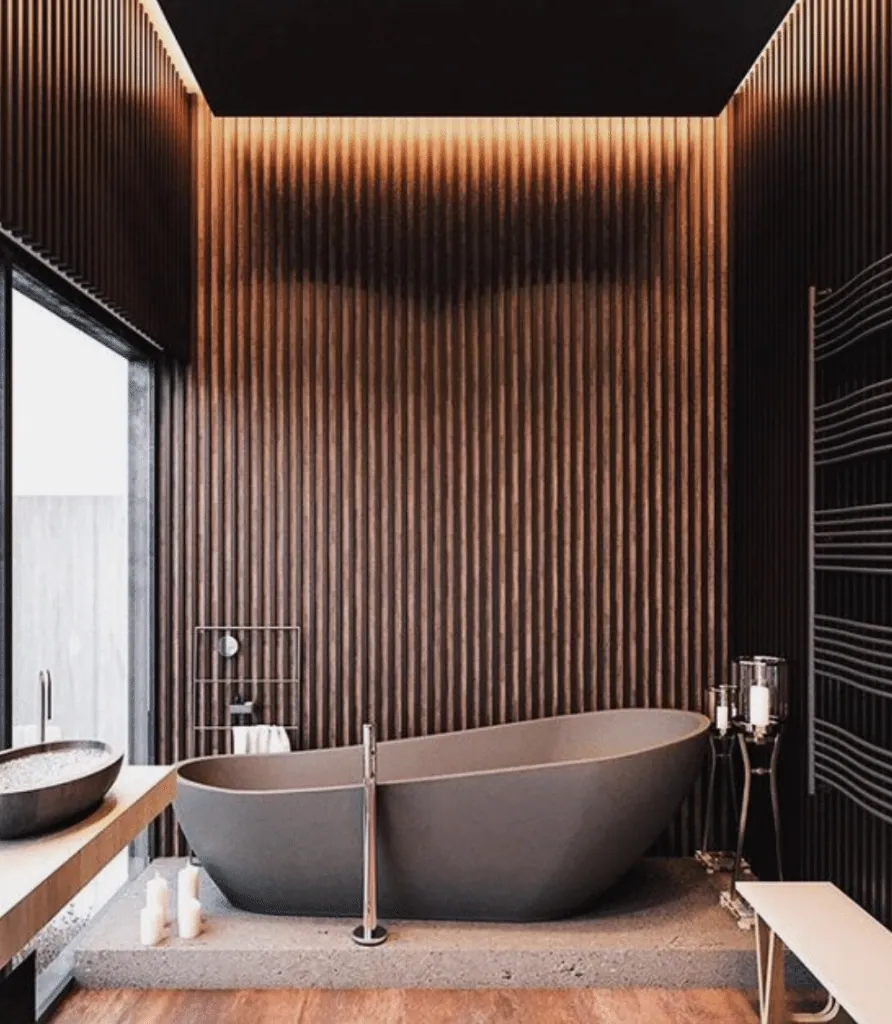 Salle de bains en tasseaux de bois sombre, lumière rasante et baignoire îlot grise sur socle
